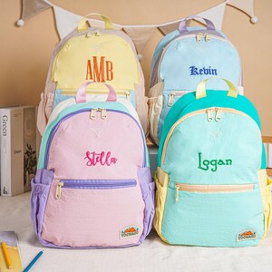 May include: Four pastel-coloured backpacks with embroidered names. The backpacks are in shades of yellow, blue, pink, and teal. Each backpack features a different name: "AMB", "Kevin", "Stella", and "Logan".