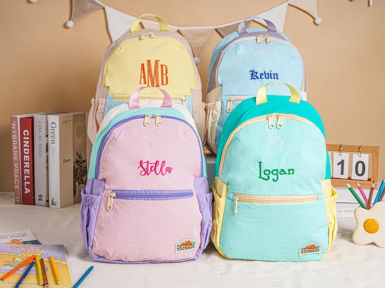 May include: Four pastel-coloured backpacks with embroidered names. The backpacks are in shades of yellow, blue, pink, and teal. Each backpack features a different name: "AMB", "Kevin", "Stella", and "Logan".