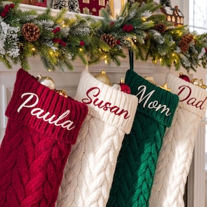May include: Four cable-knit Christmas stockings hanging in a row. The first is red with white embroidery reading Paula, the second is white with red embroidery reading Susan, the third is green with white embroidery reading Mom, and the fourth is white with red embroidery reading Dad.