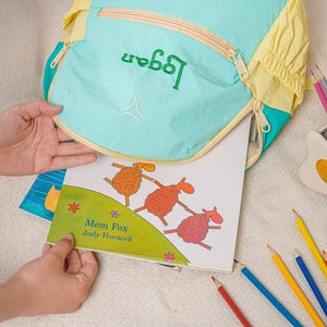 May include: A turquoise and yellow backpack with the name "Logan" embroidered in green. A children's book with illustrations of sheep is being placed inside the backpack. Coloured pencils and a pencil holder are also visible.