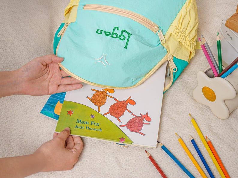 May include: A turquoise and yellow backpack with the name "Logan" embroidered in green. A children's book with illustrations of sheep is being placed inside the backpack. Coloured pencils and a pencil holder are also visible.