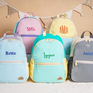 May include: Five personalised backpacks in pastel shades: light blue, pink, yellow, teal, and grey. Each backpack has a name embroidered on the front: Stella, Kevin, Logan, Josiah, and AMB. The backpacks have a front pocket and a top handle.
