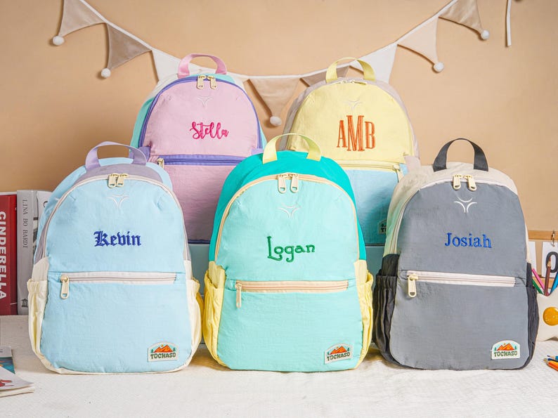 May include: Five personalised backpacks in pastel shades: light blue, pink, yellow, teal, and grey. Each backpack has a name embroidered on the front: Stella, Kevin, Logan, Josiah, and AMB. The backpacks have a front pocket and a top handle.