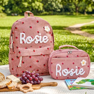 以下が含まれることがあります： イチゴ柄と白い文字で「Rosie」と書かれたピンク色のコーデュロイバックパックとランチバッグ。各バッグには白い花が飾られています。ピクニックブランケットの上にセットが置かれています。