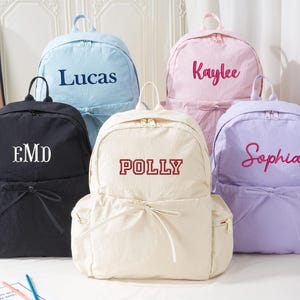 Pode incluir: Cinco mochilas personalizadas em várias cores: preto, azul claro, rosa, creme e lavanda. Cada mochila apresenta um nome ou iniciais bordados em uma cor contrastante. As mochilas têm um fecho de cordão e uma alça superior.