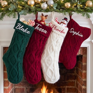 May include: Four cable-knit Christmas stockings in dark green, deep red, white, and burgundy. Each stocking features a white embroidered name: Blake, Tessa, Phoebe, and Sophia. The stockings are filled with small toys and candy canes.