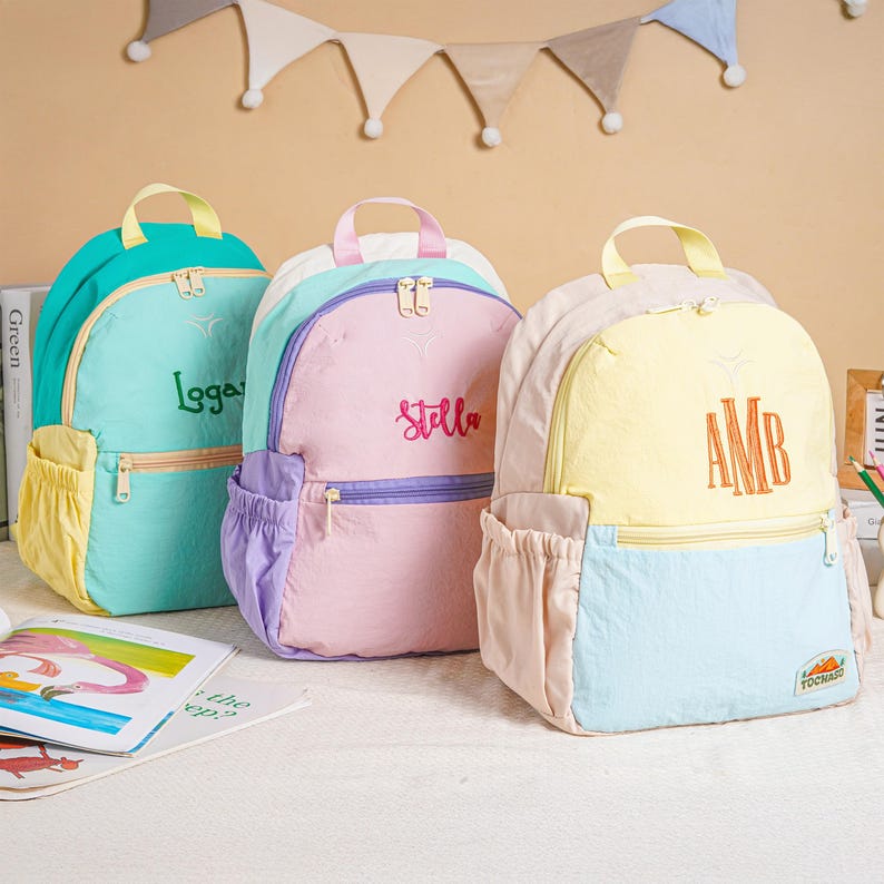 May include: Three colourful backpacks in teal, pink, and yellow. Each backpack has a top handle, a front pocket, and a zip closure. The teal backpack has the name "Logga" embroidered on it, the pink backpack has "Stella", and the yellow backpack has "AMB".