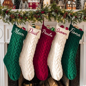 Könnte beinhalten: Fünf Weihnachtssocken aus Zopfstrick hängen in einer Reihe. Die Socken haben abwechselnde Farben in Dunkelgrün, Creme und Dunkelrot. Jede Socke hat einen umgeschlagenen Bund mit einem in Schreibschrift gestickten Namen: Mom, Dad, Paula, Susan, Fiona.