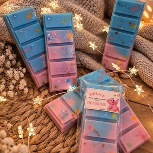 Peut inclure: Plusieurs cires parfumées rectangulaires dans des tons bleu et rose, avec des taches colorées et des paillettes. Une cire est emballée dans du plastique transparent avec une étiquette DELICE BONBONS et un ours en peluche rose.