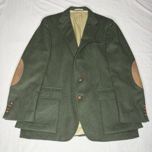 May include: A dark olive green blazer featuring tan suede elbow patches, two front buttons, two flap pockets, and a chest pocket. The sleeves have four small buttons at the cuffs, and the interior is lined with a light beige fabric.