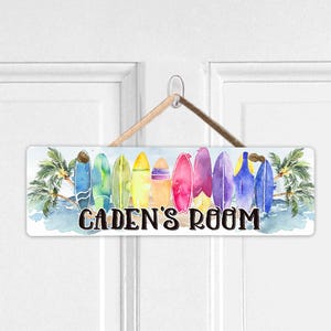 May include: A rectangular door sign with a watercolor design of surfboards in various colors, with palm trees on each end. The sign reads "CADEN'S ROOM" in black letters and is hanging from a wooden dowel.