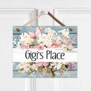 May include: A light blue wooden sign with a floral design and the text "Gigi's Place" in black lettering. The sign is hanging on a white door.