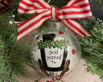 May include: Clear glass ornament with a hand-painted design that says "Just Married". The ornament has a red and white striped bow and a silver wire hanger. The ornament is decorated with red hearts and greenery.