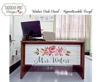 May include: A white woodgrain teacher desk decal with pink floral accents and the text "Mrs. Walters". The decal is applied to a brown wooden desk.