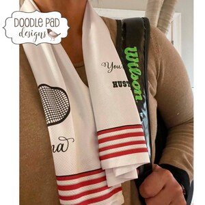 K&ouml;nnte beinhalten: Wei&szlig;es Tennishandtuch mit roten Streifen und einem schwarzen und wei&szlig;en Tennisschl&auml;ger-Motiv. Das Handtuch tr&auml;gt den Text "You're a Hustler" und "Wilson" aufgedruckt.