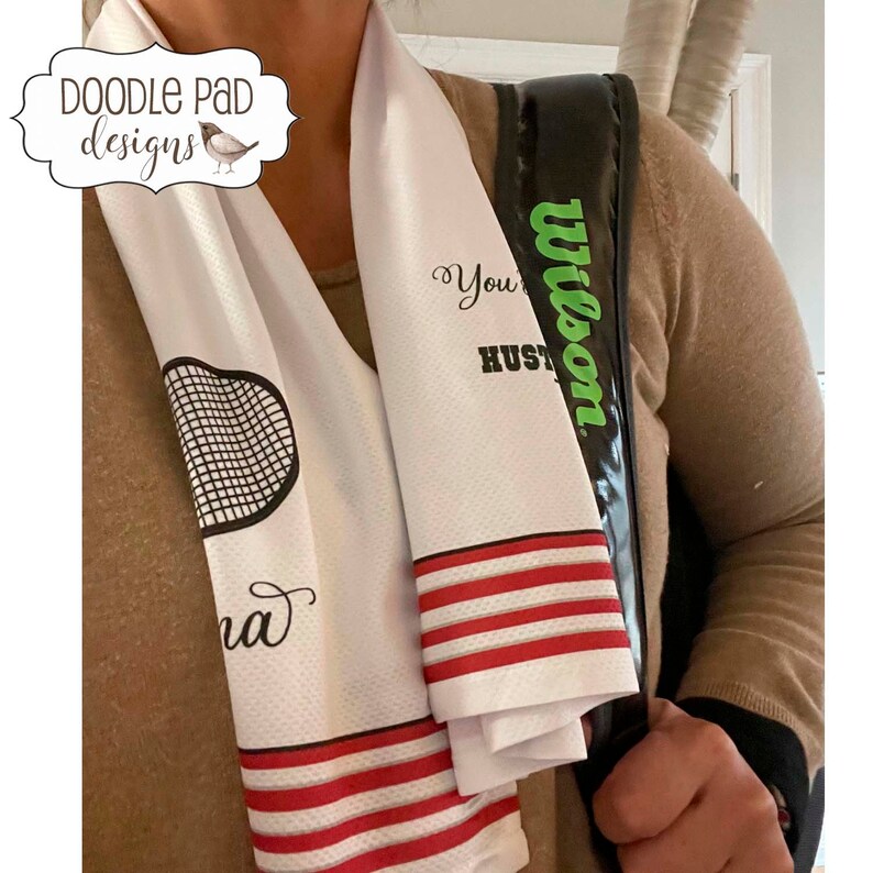 K&ouml;nnte beinhalten: Wei&szlig;es Tennishandtuch mit roten Streifen und einem schwarzen und wei&szlig;en Tennisschl&auml;ger-Motiv. Das Handtuch tr&auml;gt den Text "You're a Hustler" und "Wilson" aufgedruckt.