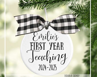 May include: A white, round ornament with the text "Emilie's FIRST YEAR Teaching 2024-2025" in black script. It is adorned with a black and white checkered bow and hangs from a Christmas tree branch. The ornament is double-sided.