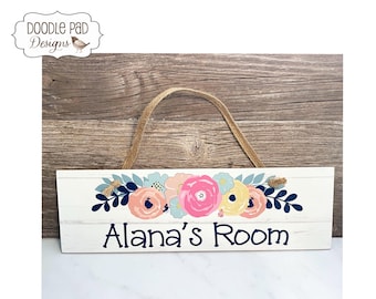 May include: A rectangular white wooden sign with a horizontal plank design, featuring a floral arrangement in shades of pink, yellow, and blue above the dark blue text "Alana's Room". The sign includes a natural jute twine hanging cord.