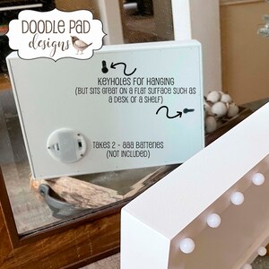 May include: White wooden sign with a white background and black text. The sign has a battery compartment on the back and keyholes for hanging. The text reads "KEYHOLES FOR HANGING (BUT SITS GREAT ON A FLAT SURFACE SUCH AS A DESK OR A SHELF) Takes 2 - aaa Batteries (NOT INCLUDED)"
