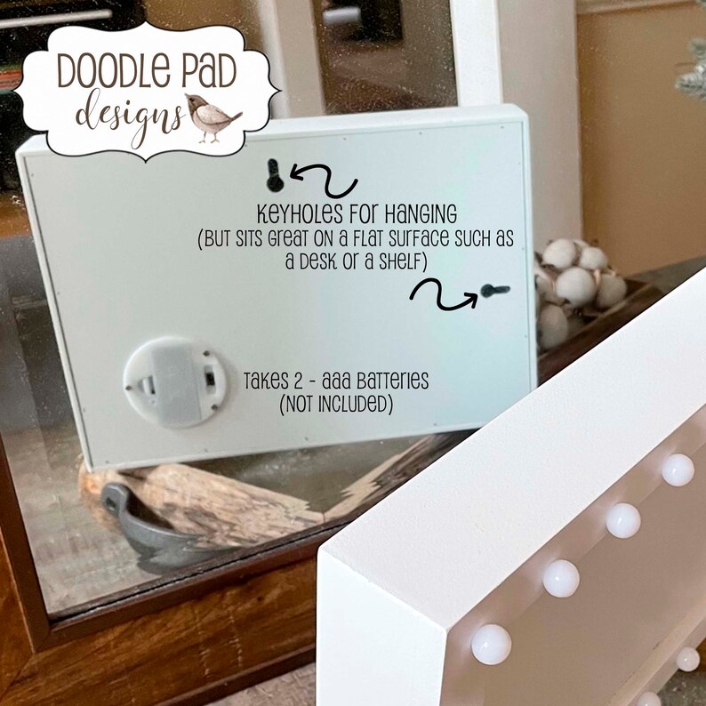 May include: White wooden sign with a white background and black text. The sign has a battery compartment on the back and keyholes for hanging. The text reads "KEYHOLES FOR HANGING (BUT SITS GREAT ON A FLAT SURFACE SUCH AS A DESK OR A SHELF) Takes 2 - aaa Batteries (NOT INCLUDED)"