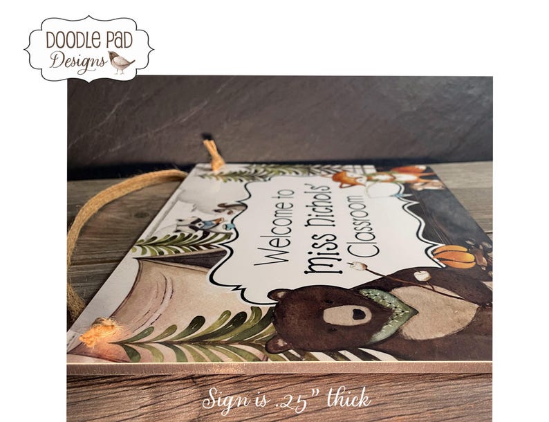 May include: A wooden classroom sign with the text "Welcome to Miss Nichols' Classroom" in a decorative font. The sign features illustrations of woodland animals and foliage. The sign is .25 inches thick and has a jute rope for hanging.
