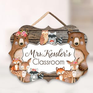 May include: Wooden classroom sign with a rustic wood grain background. The sign features a decorative border with woodland animals, including bears, deer, foxes, an owl, and a raccoon. The text "Mrs. Kessler's Classroom" is displayed in a decorative font.
