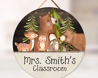 May include: A round wooden wall sign featuring a watercolor illustration of a deer, a bear, an owl, a rabbit, and a squirrel in a forest setting. The lower portion is cream-colored with the black text "Mrs. Smith's Classroom" printed on it.