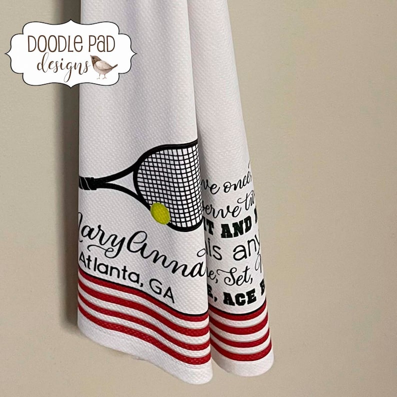 K&ouml;nnte beinhalten: Wei&szlig;es K&uuml;chentuch mit roten Streifen und einem schwarzen und wei&szlig;en Tennisracket-Motiv. Der Text "Larry Anna Atlanta, GA" und "We once serve the...it and...is any...Ace" ist auf das Handtuch gedruckt.