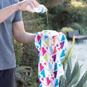 K&ouml;nnte beinhalten: Eine Person gie&szlig;t Wasser aus einer durchsichtigen Plastikwasserflasche auf ein wei&szlig;es K&uuml;hltuch mit einem bunten geometrischen Muster. Das Tuch besteht aus Mikrofaser und ist so konzipiert, dass es beim Nassen k&uuml;hl bleibt.