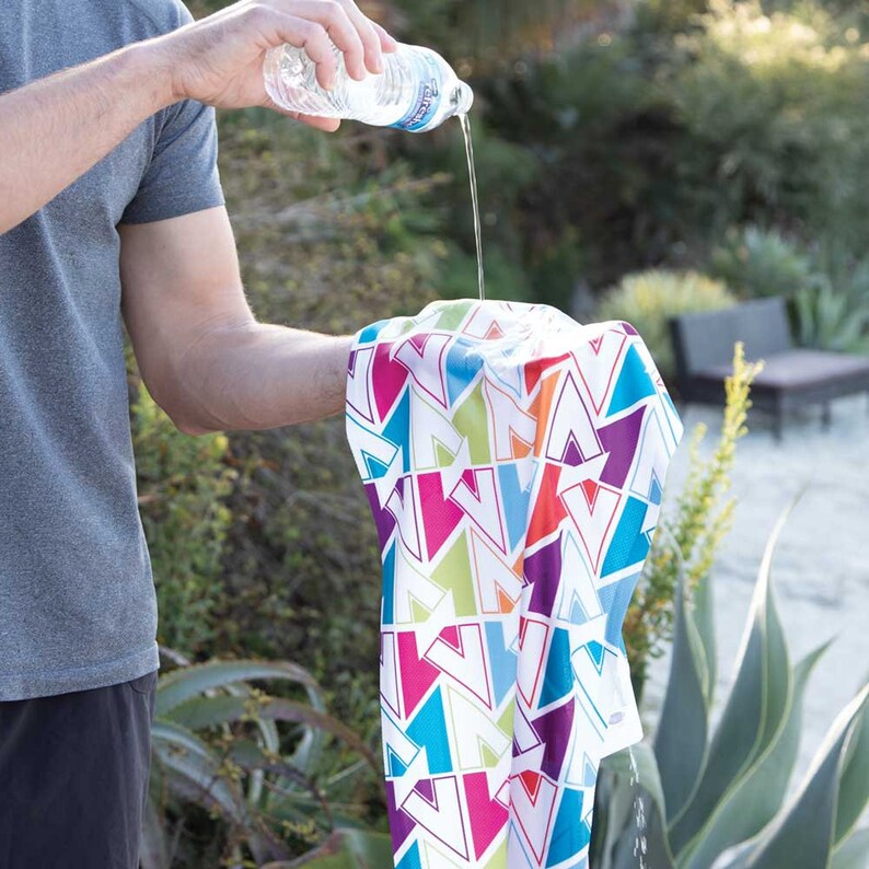 K&ouml;nnte beinhalten: Eine Person gie&szlig;t Wasser aus einer durchsichtigen Plastikwasserflasche auf ein wei&szlig;es K&uuml;hltuch mit einem bunten geometrischen Muster. Das Tuch besteht aus Mikrofaser und ist so konzipiert, dass es beim Nassen k&uuml;hl bleibt.