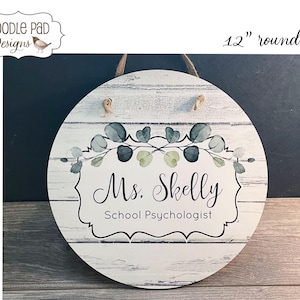 May include: A 12-inch round wooden sign with a whitewashed background. The sign features a wreath of green eucalyptus leaves and a white banner with the text "Ms. Skelly School Psychologist".