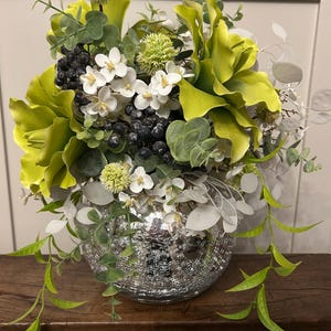 May include: A floral arrangement in a silver mercury glass vase. It features chartreuse calla lilies, small white flowers, black berries, and green and silver foliage. The vase sits on a wooden surface. The arrangement is a decorative item.