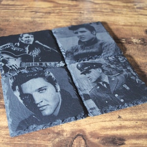 May include: Set of four square slate coasters featuring black and white images of Elvis Presley. The images depict Elvis in various poses, including playing guitar and in a military uniform. The coasters are arranged on a wooden surface.