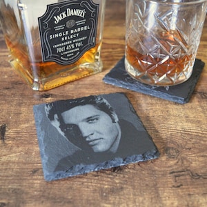 May include: A square, black slate coaster featuring a black and white portrait of Elvis Presley. A glass of amber liquid sits on a similar coaster. A bottle of Jack Daniel's Tennessee Whiskey is in the background.