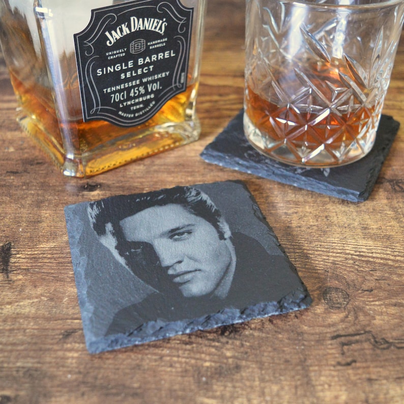 May include: A square, black slate coaster featuring a black and white portrait of Elvis Presley. A glass of amber liquid sits on a similar coaster. A bottle of Jack Daniel's Tennessee Whiskey is in the background.