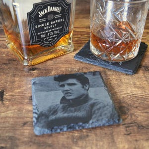 May include: A slate coaster featuring a black and white image of Elvis Presley. A bottle of Jack Daniel's Tennessee Whiskey and a glass of whiskey are also visible. The coaster is square and sits on a wooden surface.