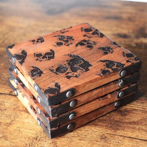 May include: Set of four wooden coasters with a skull design. The coasters are brown and have black metal accents.