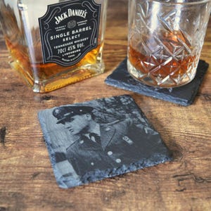 May include: A square, dark gray slate coaster with an engraved image of a person in a military uniform. A glass of amber liquid sits on a similar coaster. A bottle of Jack Daniel's Single Barrel Select whiskey is in the background.