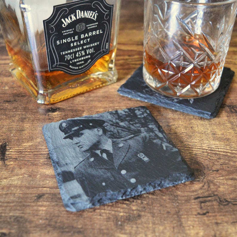 May include: A square, dark gray slate coaster with an engraved image of a person in a military uniform. A glass of amber liquid sits on a similar coaster. A bottle of Jack Daniel's Single Barrel Select whiskey is in the background.