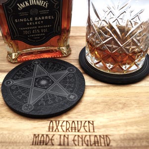 May include: A black, round coaster with a pentagram design, with a glass of whiskey on a wooden surface. A bottle of Jack Daniel's Tennessee Whiskey is in the background. The coaster has the text "AXERAVEN MADE IN ENGLAND".