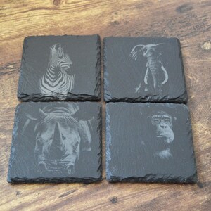 May include: Set of four square slate coasters. Each coaster features a different engraved African animal: a zebra, elephant, rhinoceros, and chimpanzee. The coasters are dark gray and rest on a wooden surface.