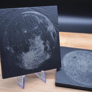 Peut inclure: Deux sous-verres en ardoise noire avec une image gravée en blanc de la lune. Les sous-verres sont posés sur une surface en bois.