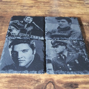 May include: Four square slate coasters featuring black and white images of Elvis Presley. The images show Elvis in various poses, including playing guitar, and in a military uniform. The coasters are arranged on a wooden surface.