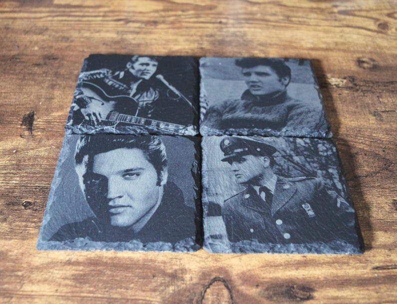 May include: Four square slate coasters featuring black and white images of Elvis Presley. The images show Elvis in various poses, including playing guitar, and in a military uniform. The coasters are arranged on a wooden surface.