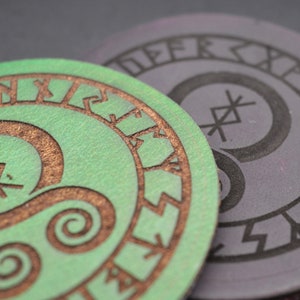 May include: Two round leather coasters in green and purple. Each coaster features a circular design with a stylised symbol in the centre and a ring of runes around the edge.