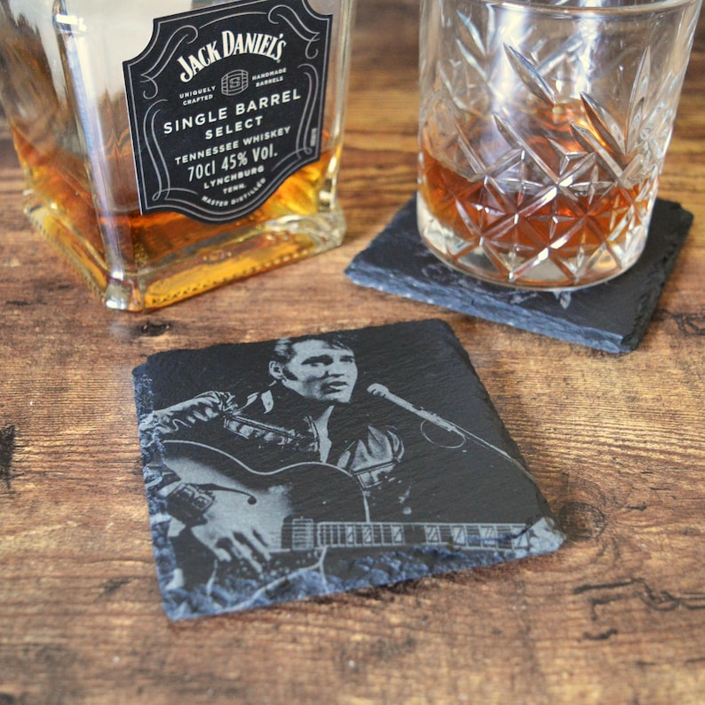 May include: Black slate coasters with a portrait of Elvis Presley playing guitar. A bottle of Jack Daniel's Single Barrel Select whiskey and a glass of amber liquid sit nearby. The coasters have a rough, natural edge.