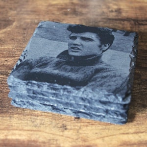 May include: A stack of four square slate coasters. Each coaster features a black and white image of a man in a sweater. The coasters have a rough, natural edge and are stacked on a wooden surface.