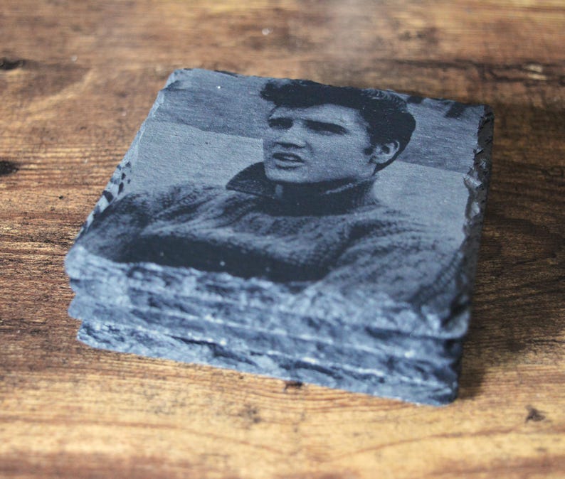 May include: A stack of four square slate coasters. Each coaster features a black and white image of a man in a sweater. The coasters have a rough, natural edge and are stacked on a wooden surface.