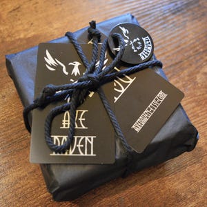 May include: A wrapped package in black paper tied with black twine. Attached are black tags with white graphics and text, including a stylized bird and the text "AXE RAVEN". The package rests on a wooden surface.