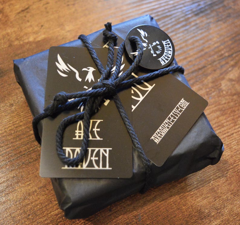 May include: A wrapped package in black paper tied with black twine. Attached are black tags with white graphics and text, including a stylized bird and the text "AXE RAVEN". The package rests on a wooden surface.